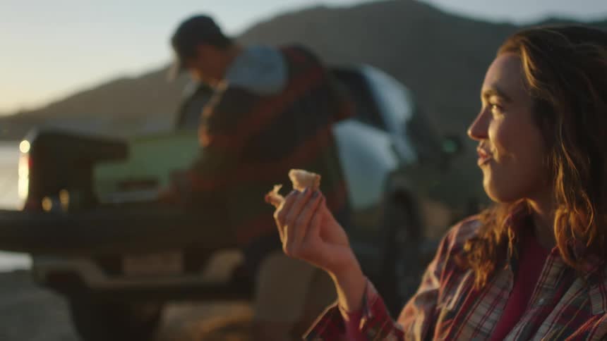 A woman eats a sandwich and laughs while a man puts something in a truck near a body of water