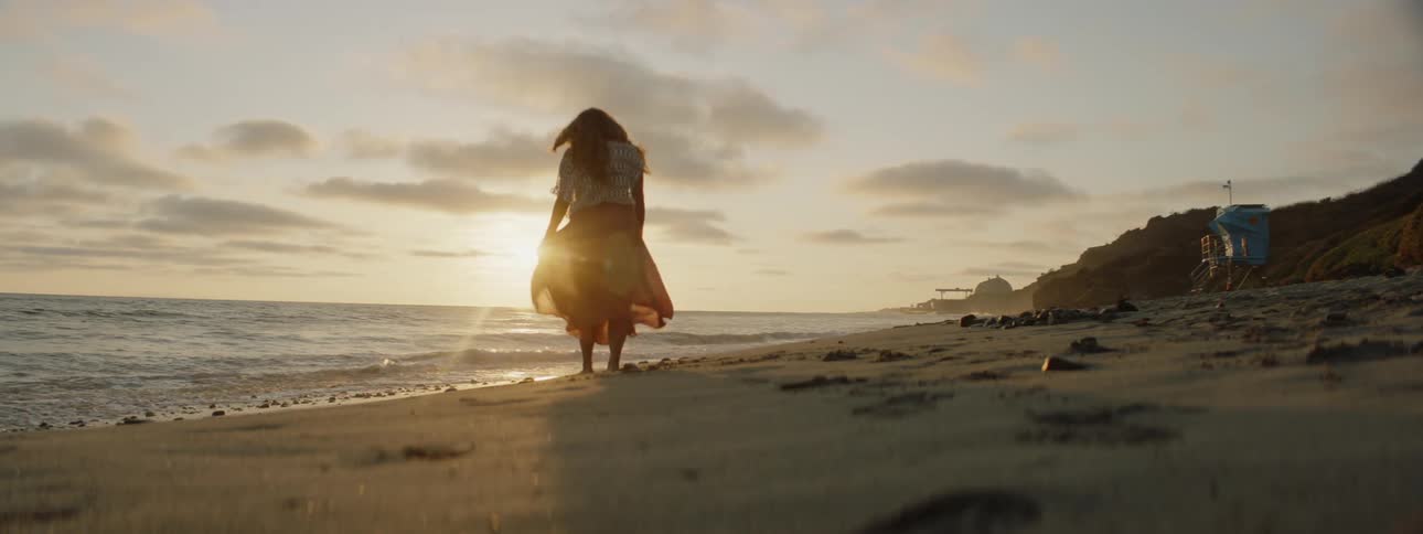 A womans long skirt blows in the wind as she walks across a beach as waves roll in to shore and the sun is on the horizon