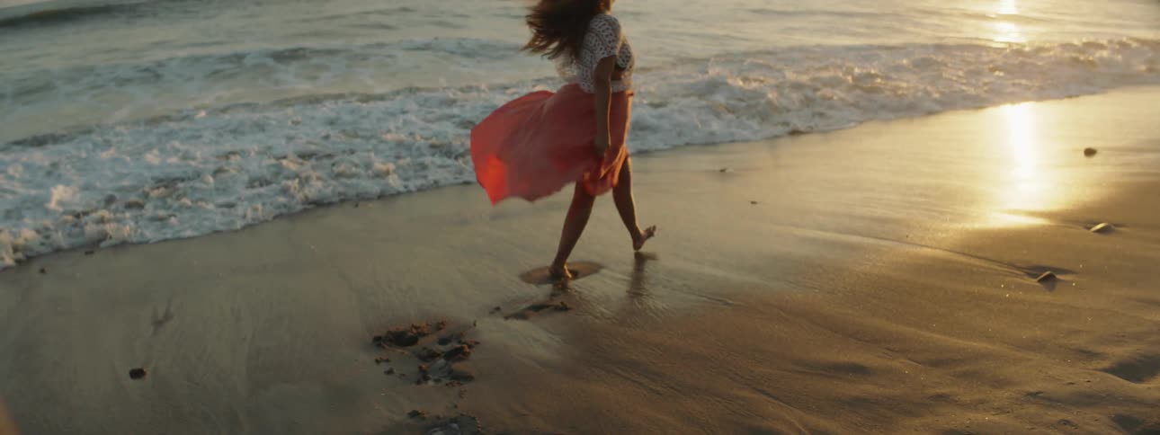 Young woman wearing a long dress walks on the sand next to the coast at sunset