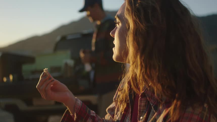 Young woman eating food cooked on a grill on the back of a truck near a beach at sunset
