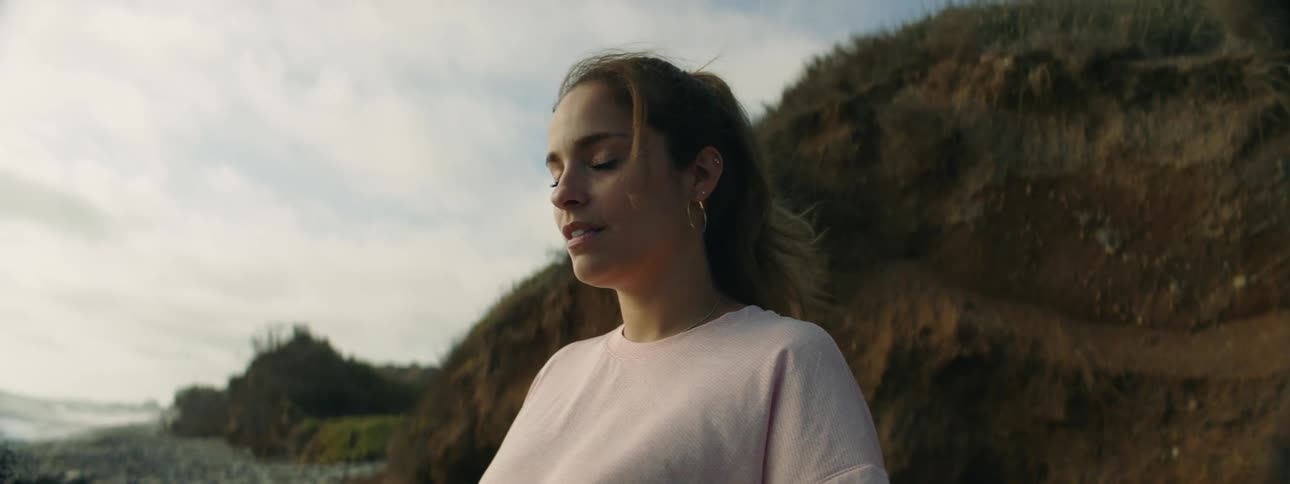A young woman stands on the shore and looks out at the water and turns her head and moves her hair off her face