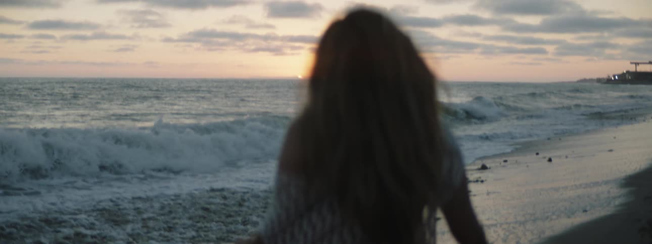 A woman is walking on a beach and her hair blows in the wind as waves roll in to shore and the sun is on the horizon