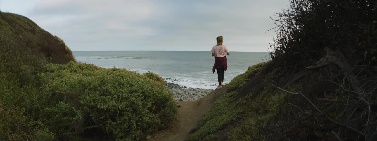 Young woman jogging on a dirt path stops to look a the ocean from a beach