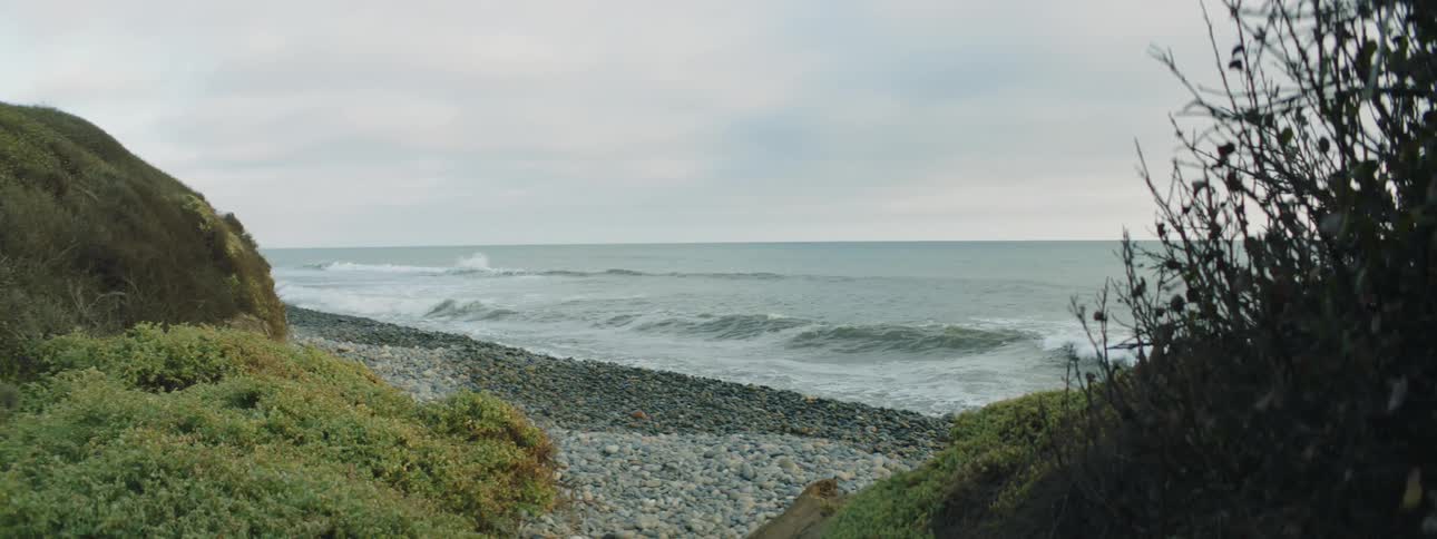Waves crash onto pebbles at shore near vegetation covered hills