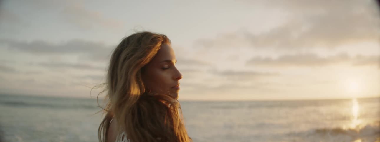 Woman with brown wavy hair and white swimsuit cover ups looks at camera smiling on the beach at sunset