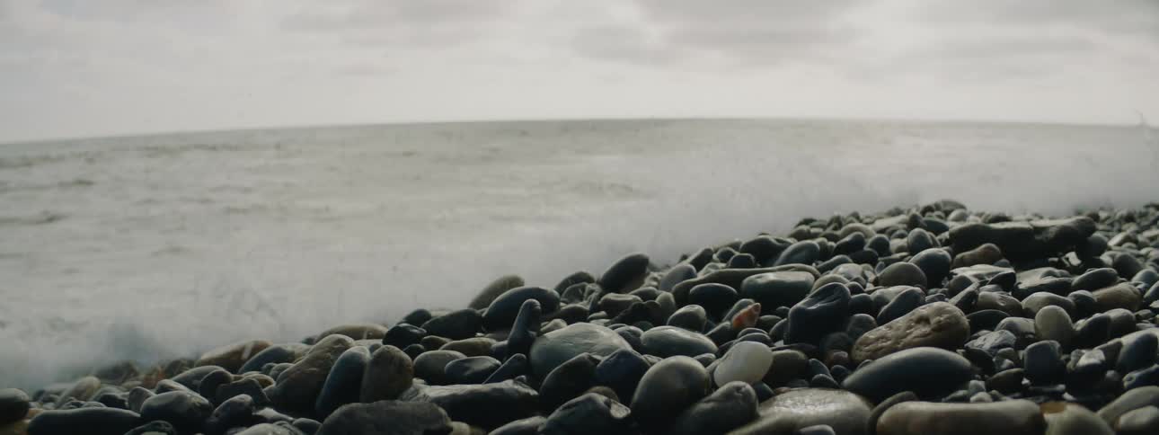 Waves crash against pebble strewn shore under an overcast sky