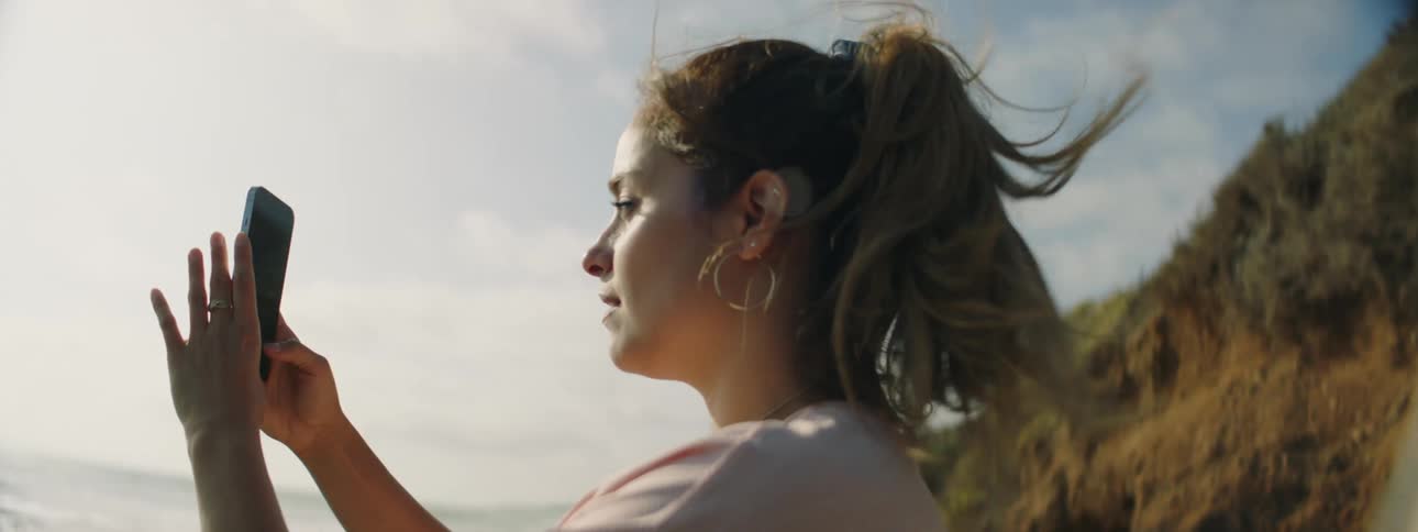 Woman with brown ponytail hoop earrings and pink t shirt takes photos on the beach next to sea