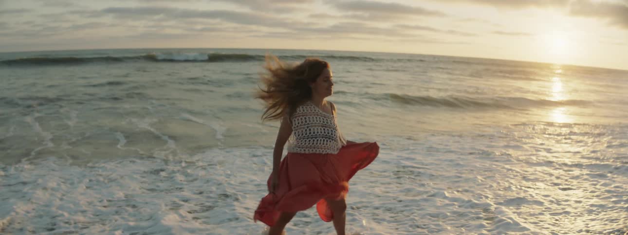 A woman holds her skirt as she walks and skips on a beach