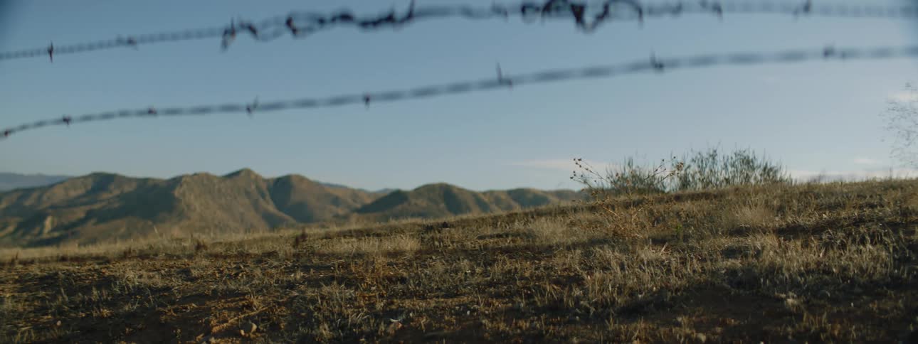 A barbed wire fence in a rural field with hills in the distance