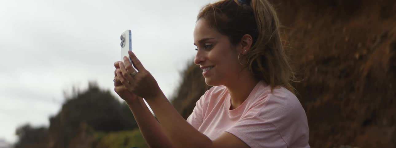 Woman with brown ponytail hoop earrings and pink t shirt takes selfie with smartphone on the coast