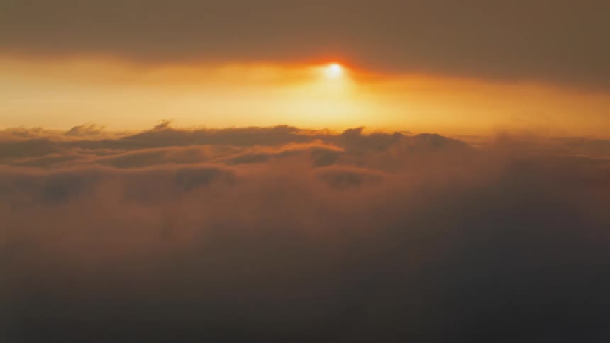 The sun shines golden as it sets above rolling clouds with a mountain peak barely visible