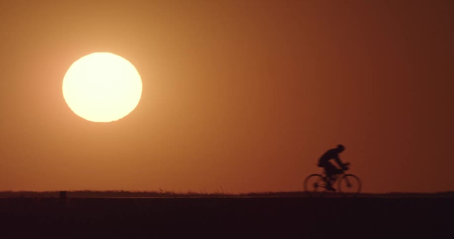 A silhouette bicyclist riding in front of a bright sun and orange sky