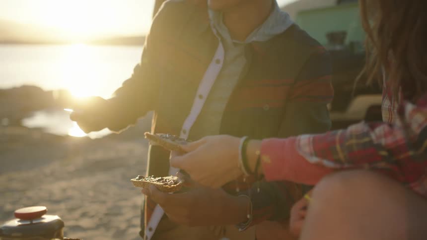 Women and men have a picnic next to black pickup truck on the beach at sunset