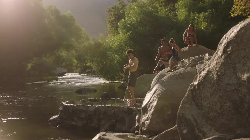 Man enters natural tub as friends climb down boulders at side of river to join him