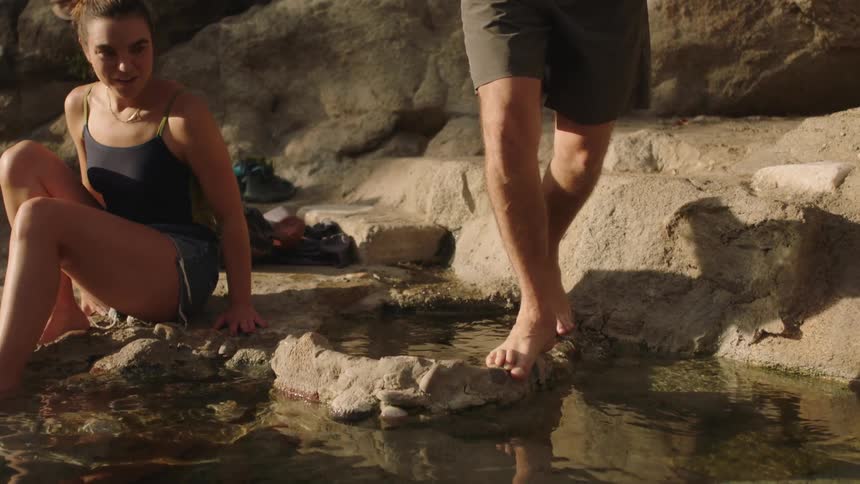 Man wades into water as woman sits on shore with her feet in water
