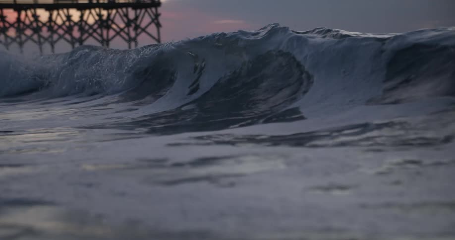 Pier is silhouetted against red sky as large wave crashes and creates foam