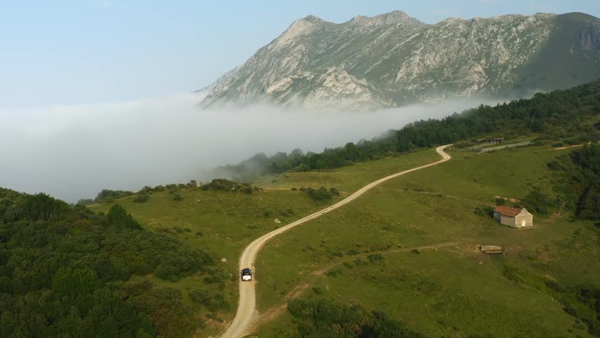 An aerial view of a car traveling on a rural dirt road with mountains and clouds on the horizon