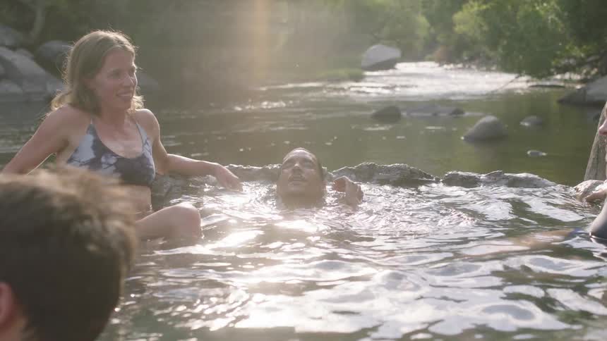 A small group of people are relaxing in a river and leaning on rocks as one man goes underwater