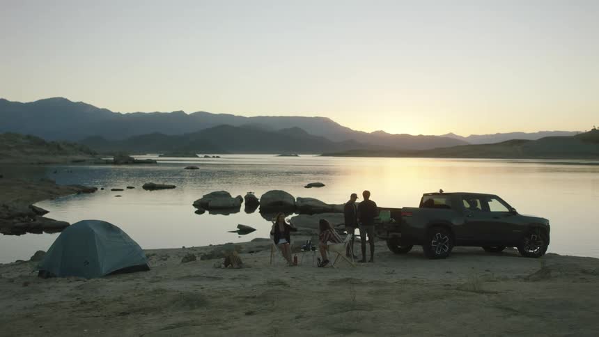 Friends camp on shore across water from sun sitting on horizon over misty mountains
