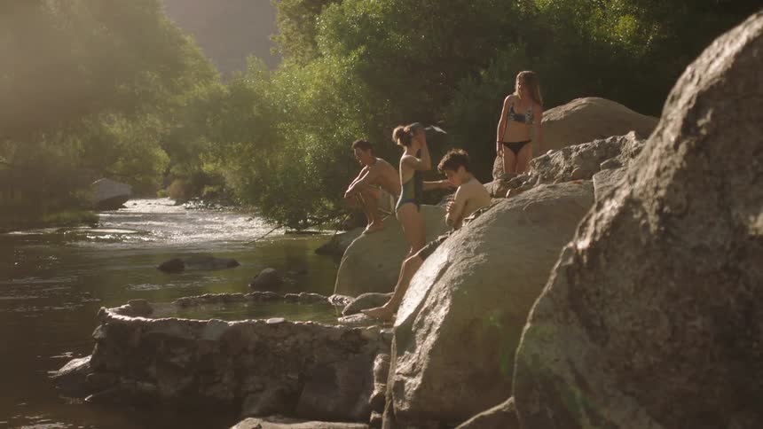 Four teens hang out on the rocks near a river while one teen throws a clothing item into the woods
