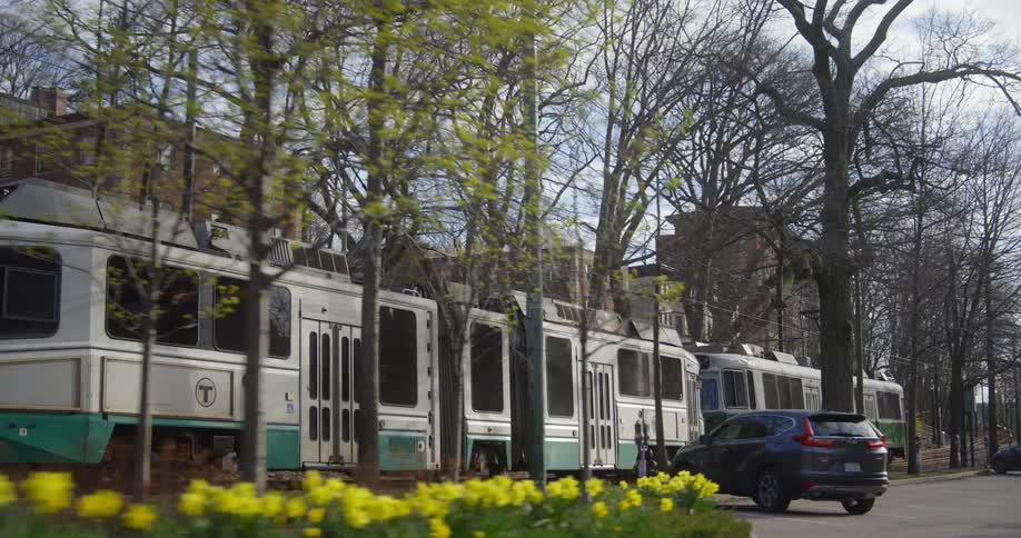A public transit train travels by buildings and a parking lot with bare trees under a cloudy sky