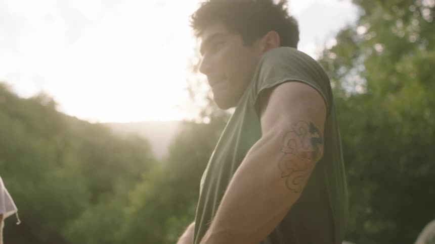 Man with green t shirt black pants takes off boots and t shirt smiling at woman standing next to him by the stream in the forest at sunset