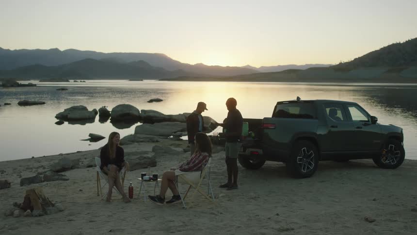 Two men standing by the truck making hot coffee while women sit talking and smiling by the lake