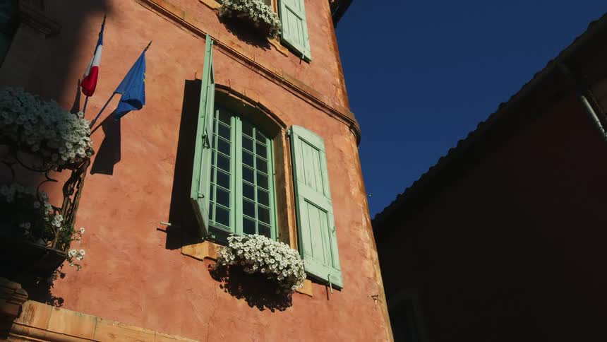 The sun shines on a building with green window trim and open shutters as two flags move in the breeze