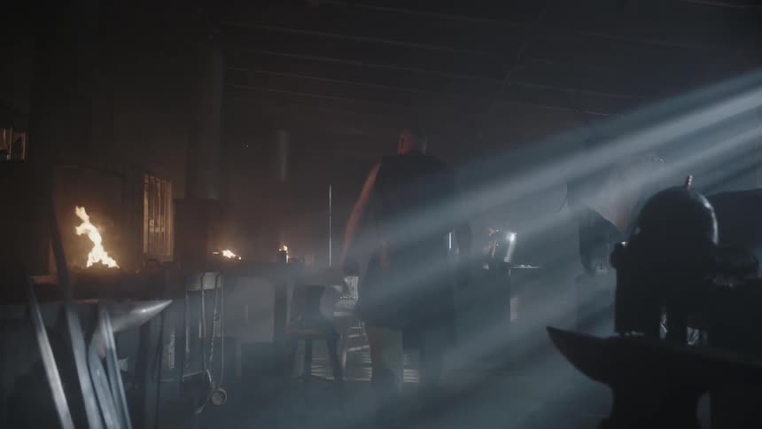 A large man walks over to a forge as sunlight streams into the workshop
