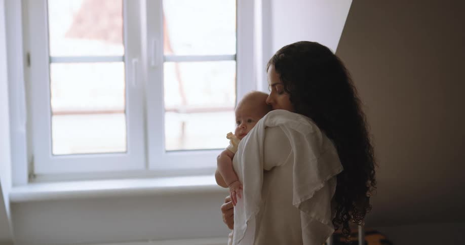 A woman cradles a baby in her arms while standing near a window indoors during the day