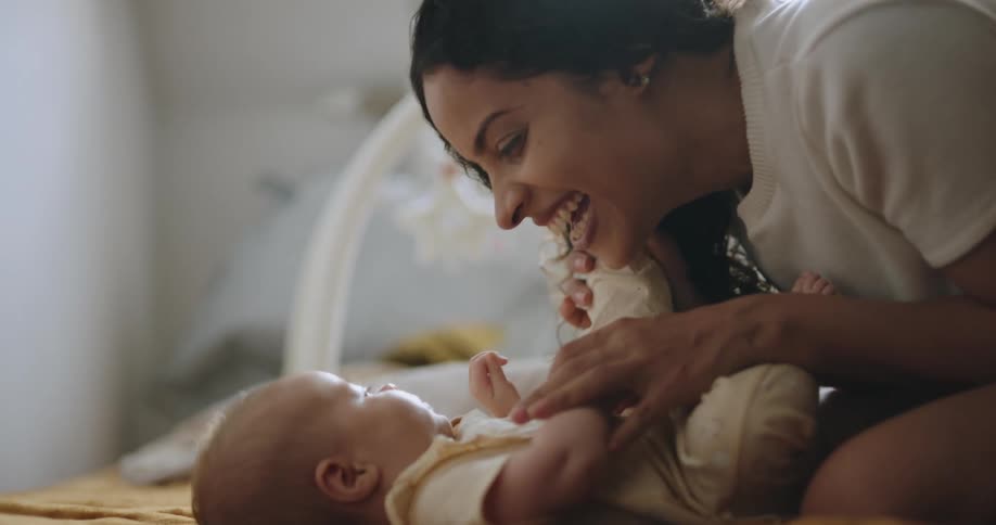 A baby is lying an a crib and their mother is leaning over holding their feet and kisses their hand as she talks and smiles