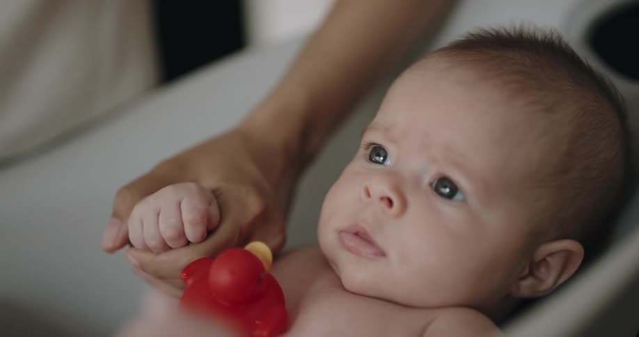 Mother holds babys hand while bathing child as red rubber ducky sits on the childs chest