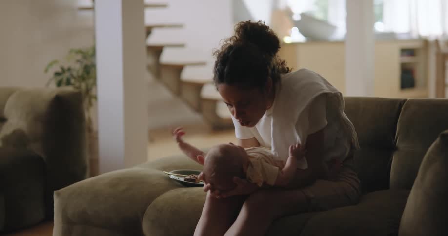 Mother with curly hair white shirt and brown shorts sits on olive couch holding newborn baby in her arms in living room