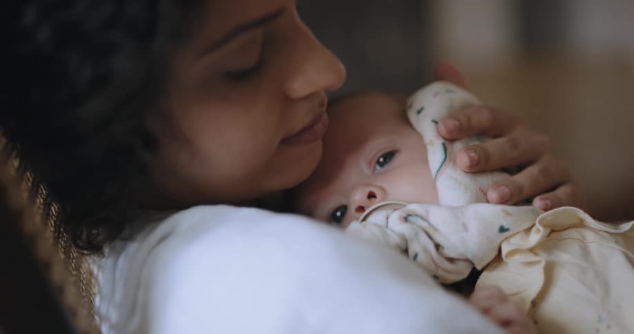 A woman is sitting in a rocking chair and holding her baby in her arms as she strokes their face with her hand