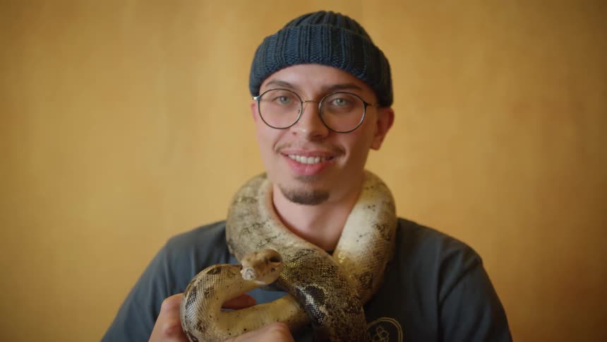 A man with a snake wrapped around his neck kisses it and laughs