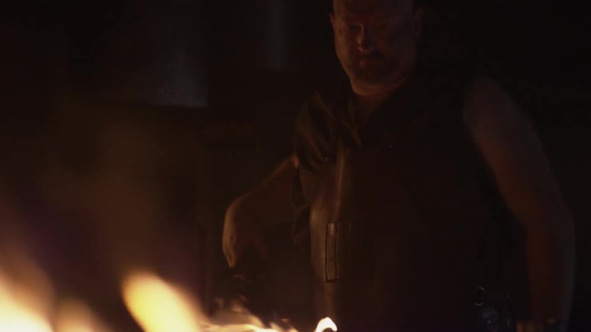 A large man tends to a tool near a fire inside a dim building