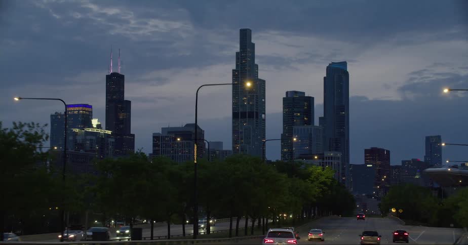 Cars traveling on an urban street with skyscrapers in the distance at dusk