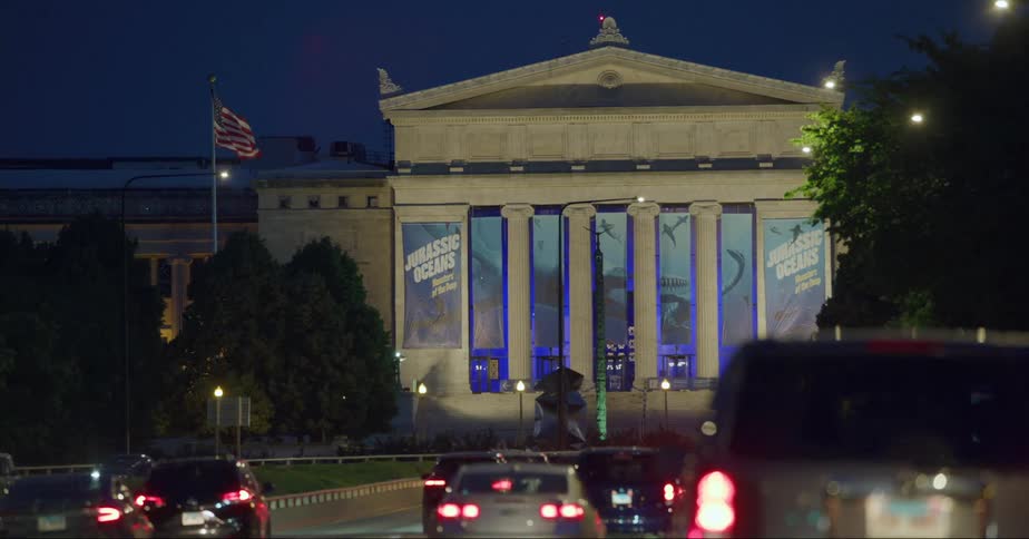 Traffic moves along streets near building with classical architectural displaying banners regarding an exhibit