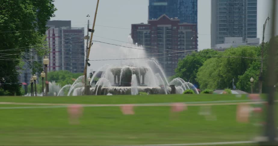 People are walking near a park with a water fountain and there are downtown buildings in the distance