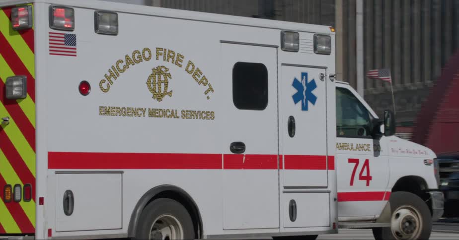 An ambulance with its lights flashing drives past people on the sidewalk in front of office buildings in a downtown area