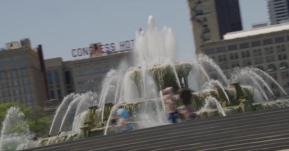 People riding bikes and cars driving past a fountain in a park in front of high rise office buildings