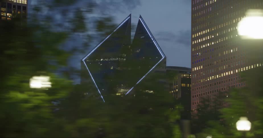 Lit streetlights and trees line road as skyscraper with geometric top stands among other downtown skyscrapers in the background