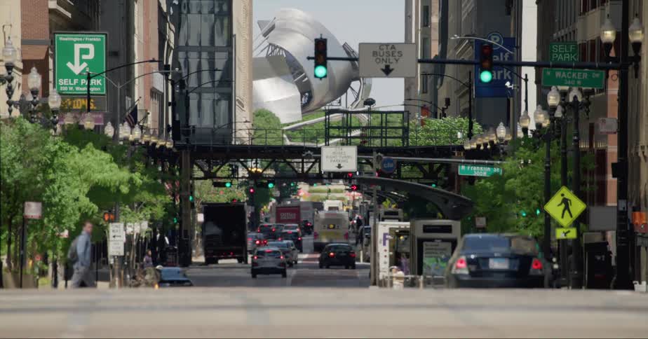 A man crosses a city street with cars driving on it as another man pushes a dolly towards a shipping truck with someone loading it