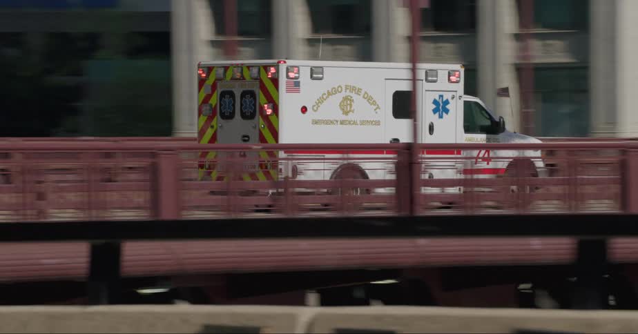 An ambulance with the flashers on travels over a bridge on a downtown street