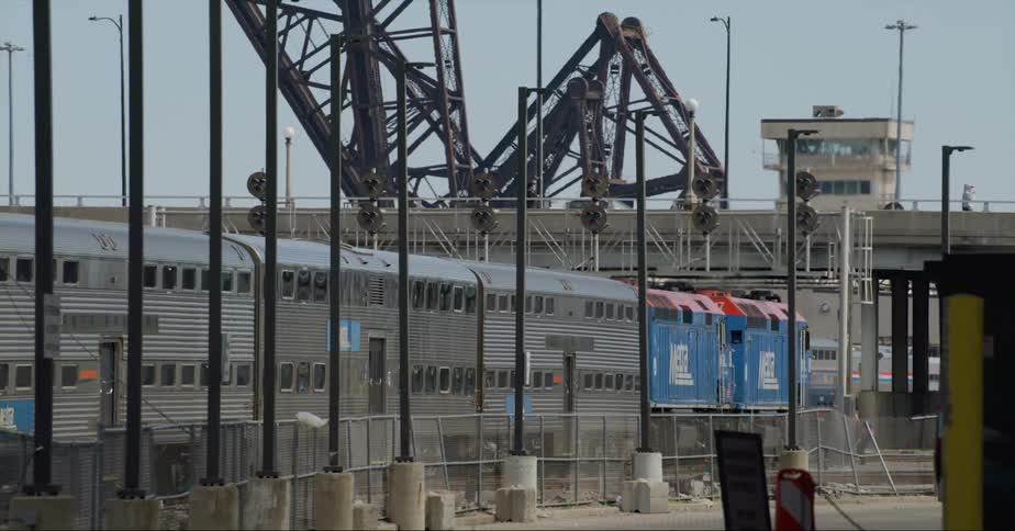 A train moves on tracks as an employee walks behind it and people jog above the train over an overpass
