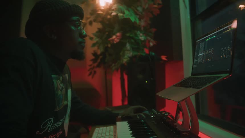 A man sitting at a desk with a laptop a musical keyboard and computer keyboard adjusts sound levels as he records