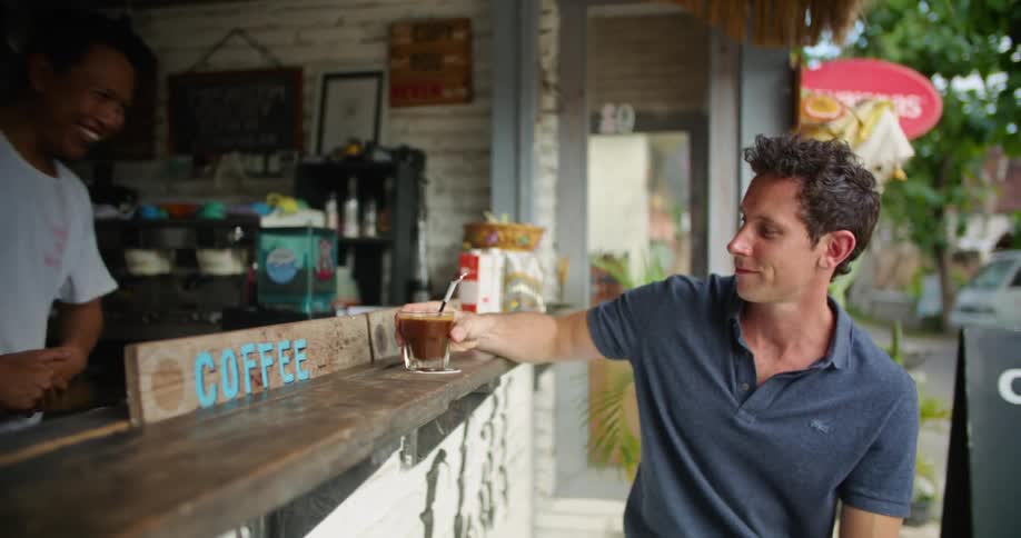 A smiling worker at an outdoors coffee bar hands a drink to a customer sitting at the bar