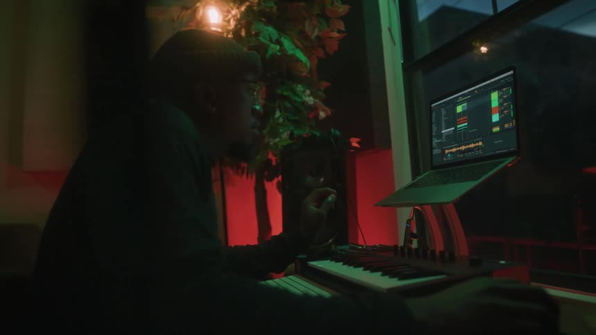 A man sitting at a desk with a laptop and musical keyboard recording and adjusting music