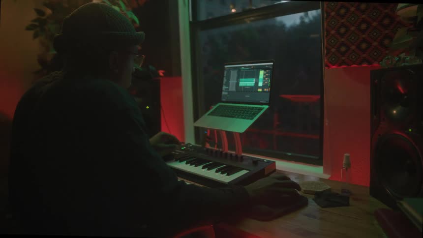 A man sits at a desk working on something music related with a laptop and a musical keyboard