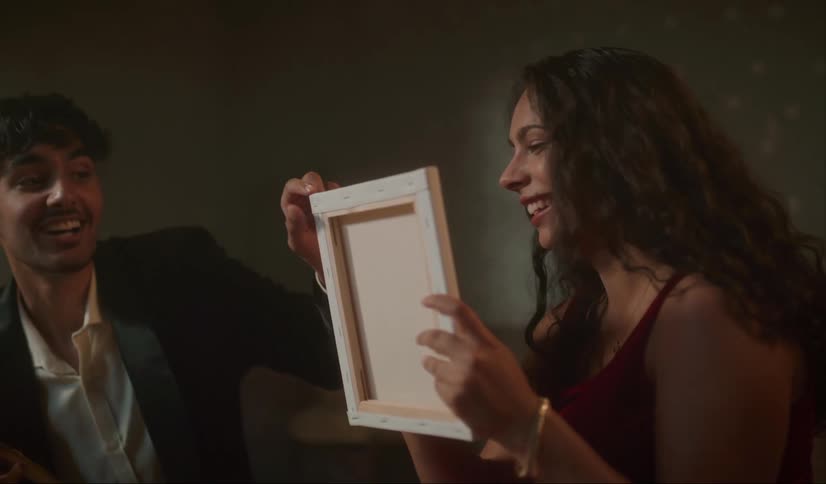 A man is holding an easel and a woman is holding a canvas as the woman paints something she doesnt want the man seeing while they both laugh